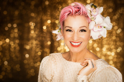 Crystal Lewis smiling with pink hair and white orchids against a gold background.