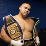 Erick Correa poses with championship belts in a blue-brick locker room.
