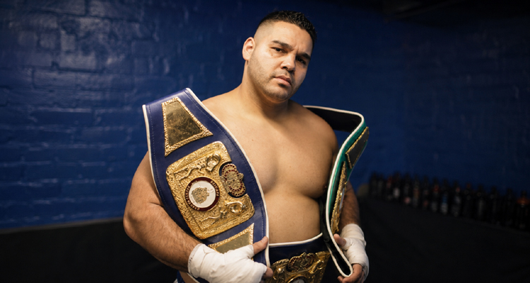 Erick Correa poses with championship belts in a blue-brick locker room.