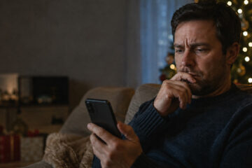 A concerned man reads an update on his smartphone beside a Christmas tree.