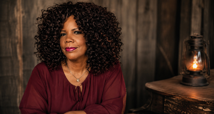 Lynda Randle seated beside a glowing lantern in a rustic setting.
