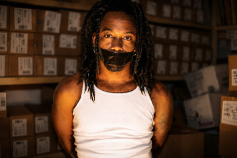 Tonéx stands in a warehouse surrounded by boxes with black tape over his mouth.